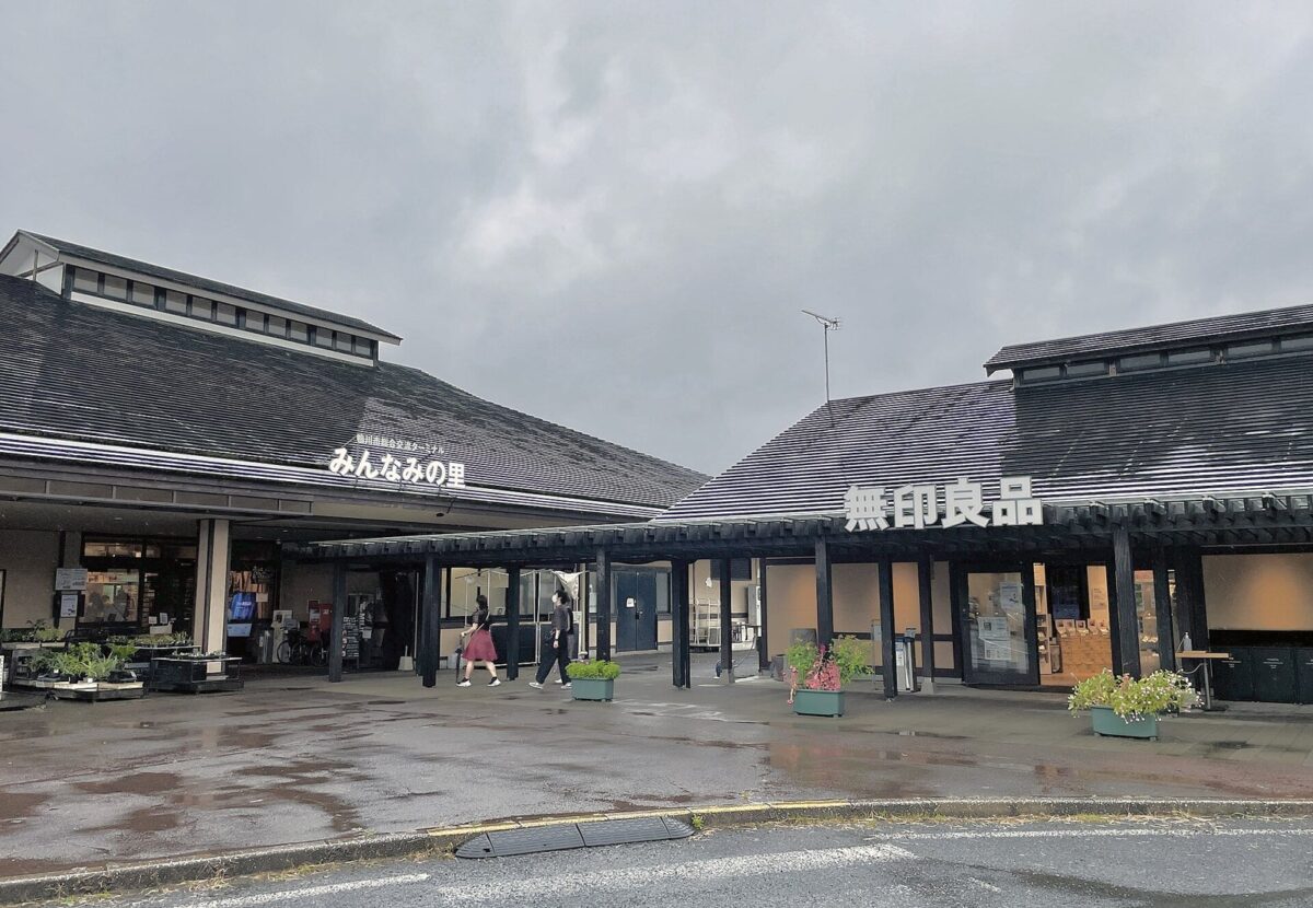 曇りの日、黒い瓦屋根の平屋が2棟並び、左の屋根に「みんなみの里」、右の屋根に「無印良品」の文字。手前に花のプランターがある