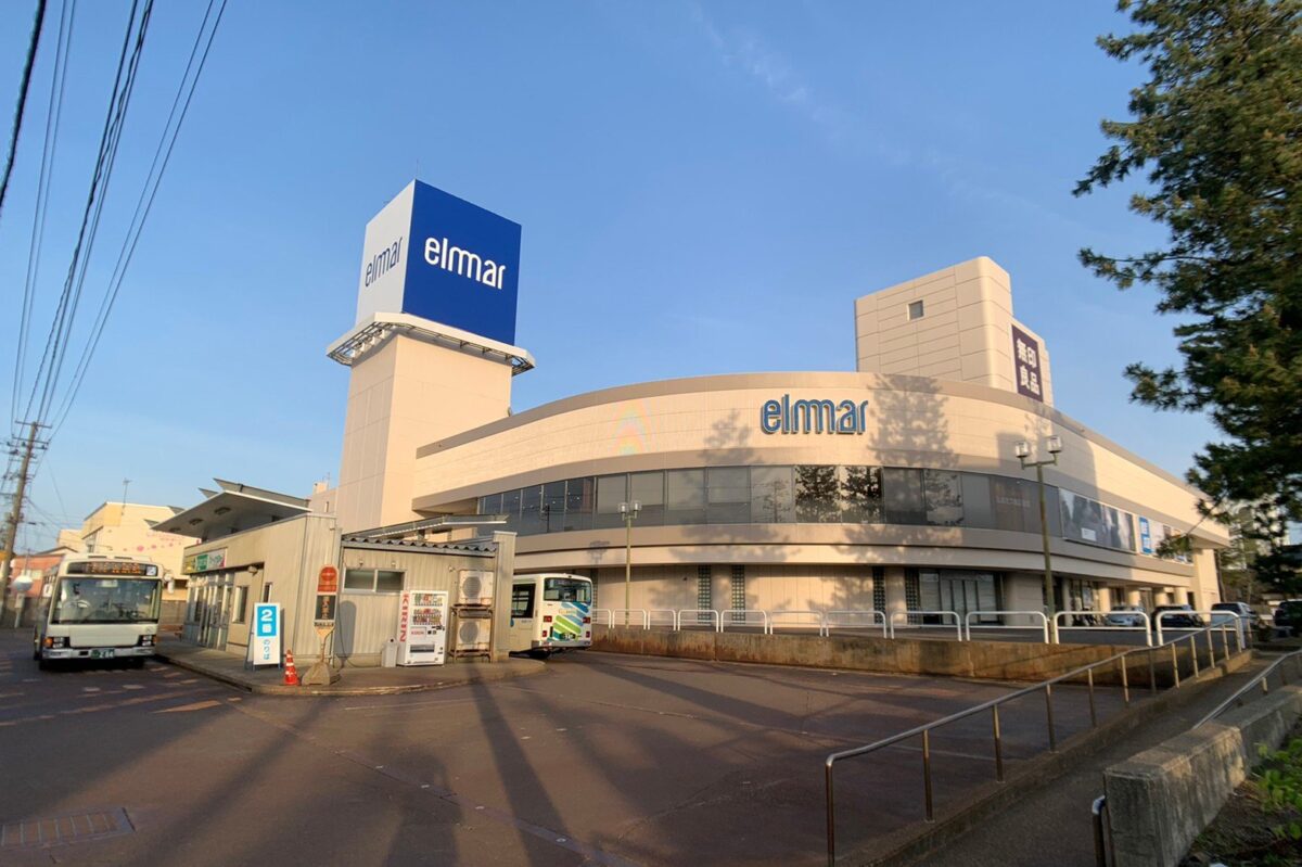晴れた夕方、バス乗り場の奥に「elmar」の大きな看板の白い建物があり、右の塔に縦書きの「無印良品」の看板が出ている