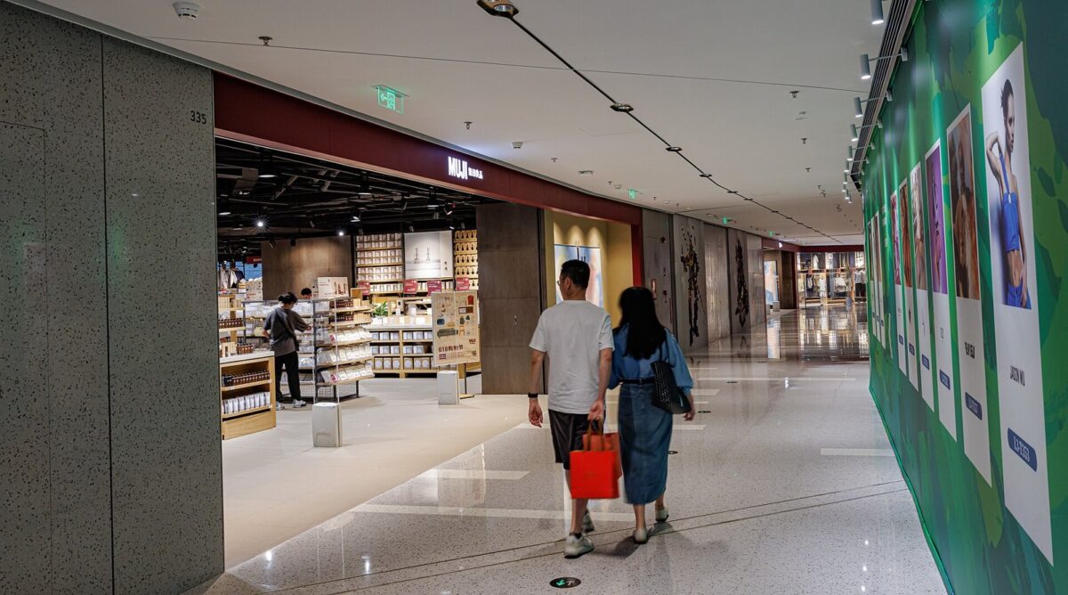 ショッピングモールの通路の左に「MUJI 無印良品」の看板の店の入口があり、木の棚に白い箱や生活雑貨が並ぶ。手前を2人が歩いている