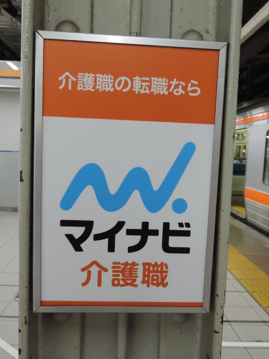 駅のホームの柱にかかった広告。オレンジの帯に「介護職の転職なら」、白地に青いマークと「マイナビ介護職」の文字