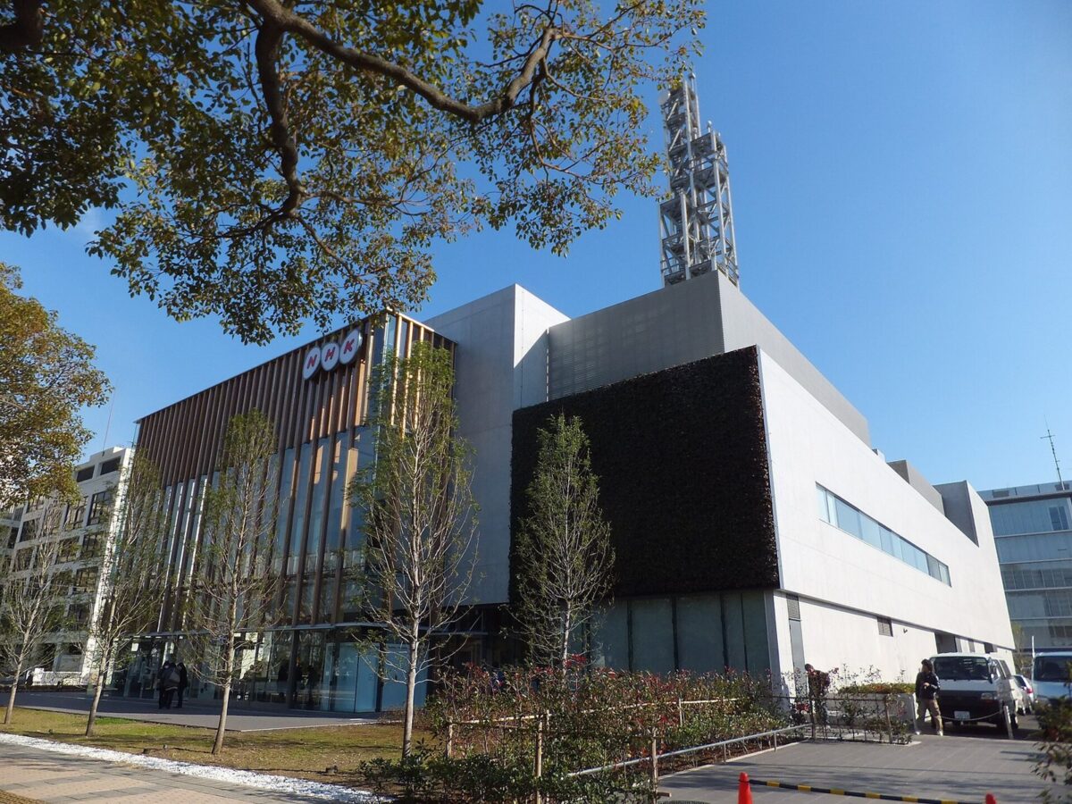 木の枝の向こうに見える、茶色と白の壁で屋上に鉄塔が立つNHK千葉放送局の建物