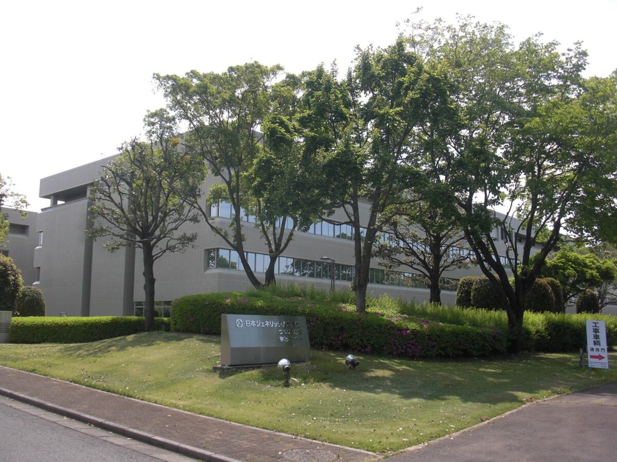 木々と芝生の奥に建つ、灰色の大きな3階建ての建物。手前の芝生に「日本ジェネリック株式会社 つくば工場」の看板が立つ