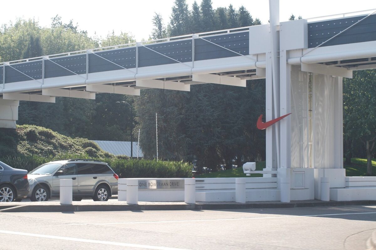 木々に囲まれた白い門。柱にオレンジ色のNikeのマークがあり、手前に車が停まっている