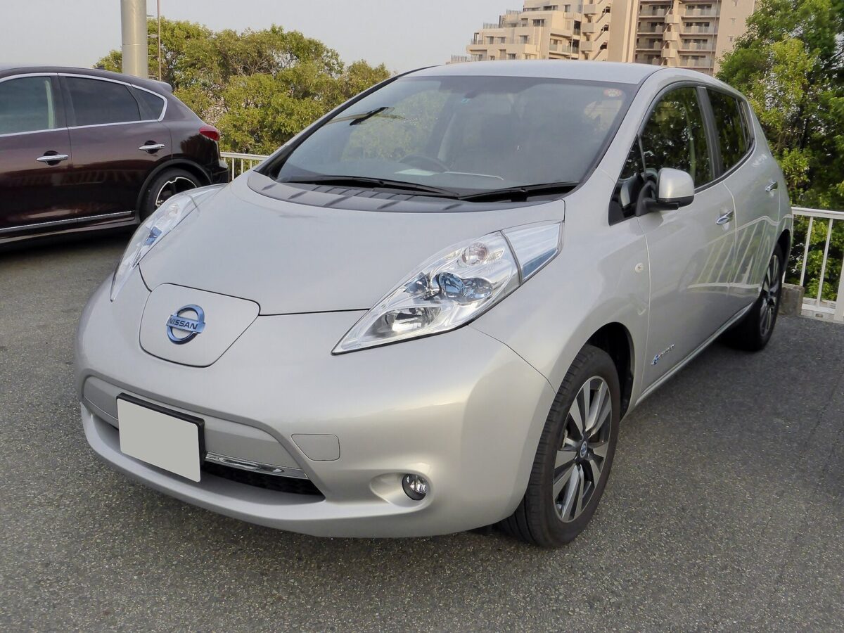 駐車場にとまる、丸みのある銀色の5人乗りの電気自動車。前に細長いライトと日産のマーク