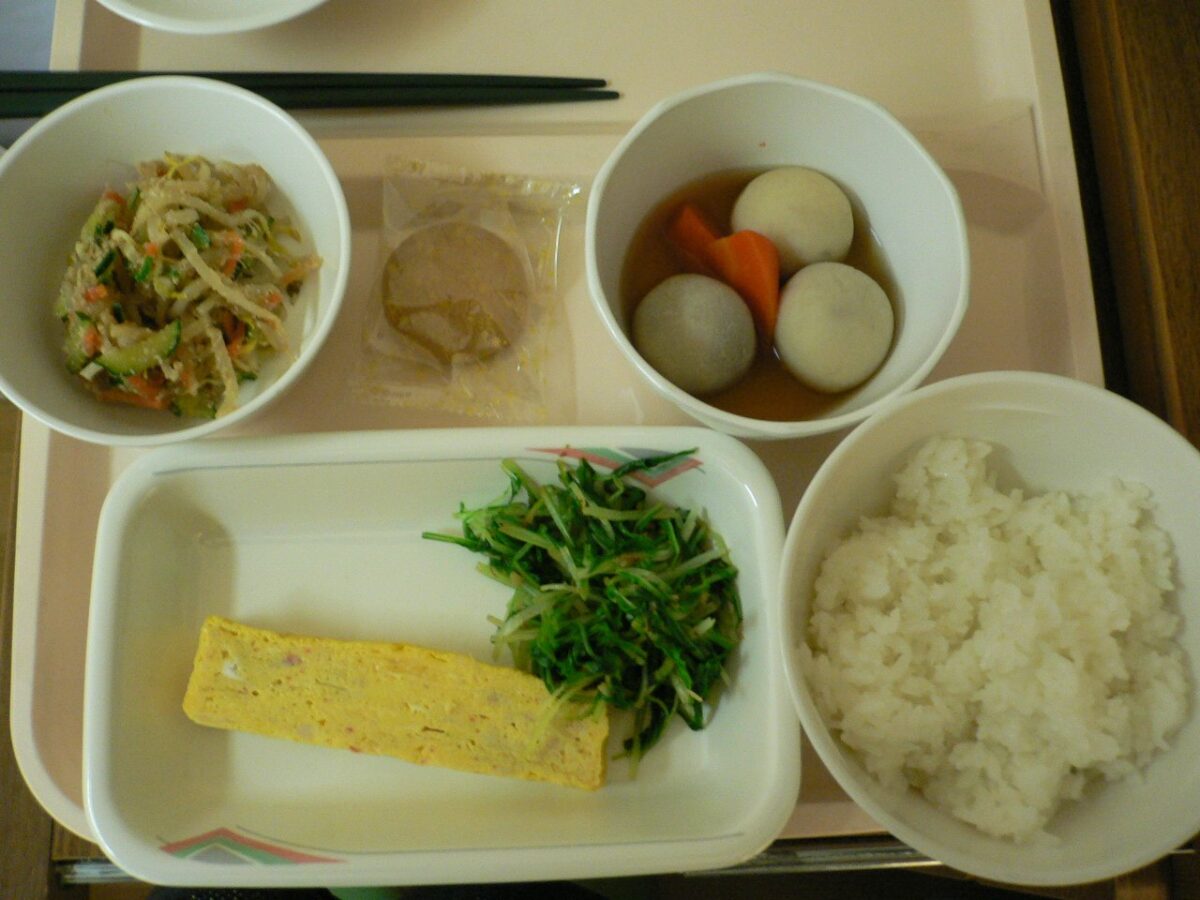 うすいピンクのトレーに、白いごはん、卵焼きと青菜の皿、里いもとにんじんの煮物、もやしのあえもの、小さな袋の佃煮が並んでいる