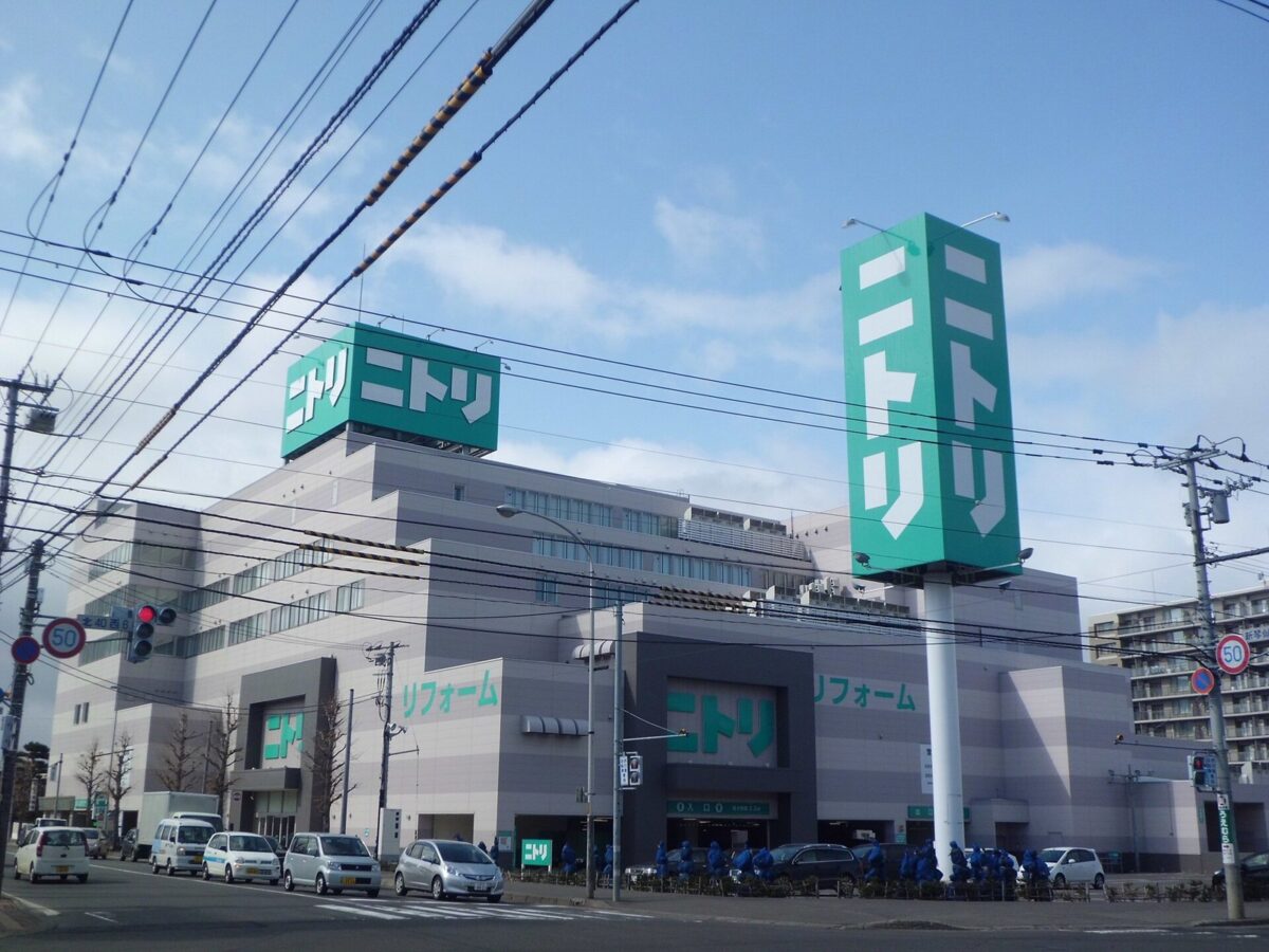 青空の下、緑の「ニトリ」の大きな看板が立つ、灰色の大きなビル。手前の駐車場に車が並ぶ