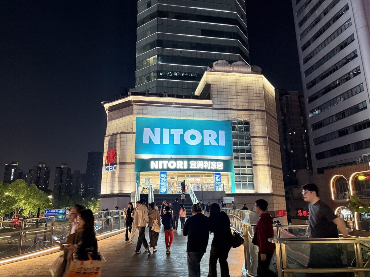 夜の高層ビルの間に、青い「NITORI」の看板を掲げた明るい建物があり、手前の広場を人が歩いている