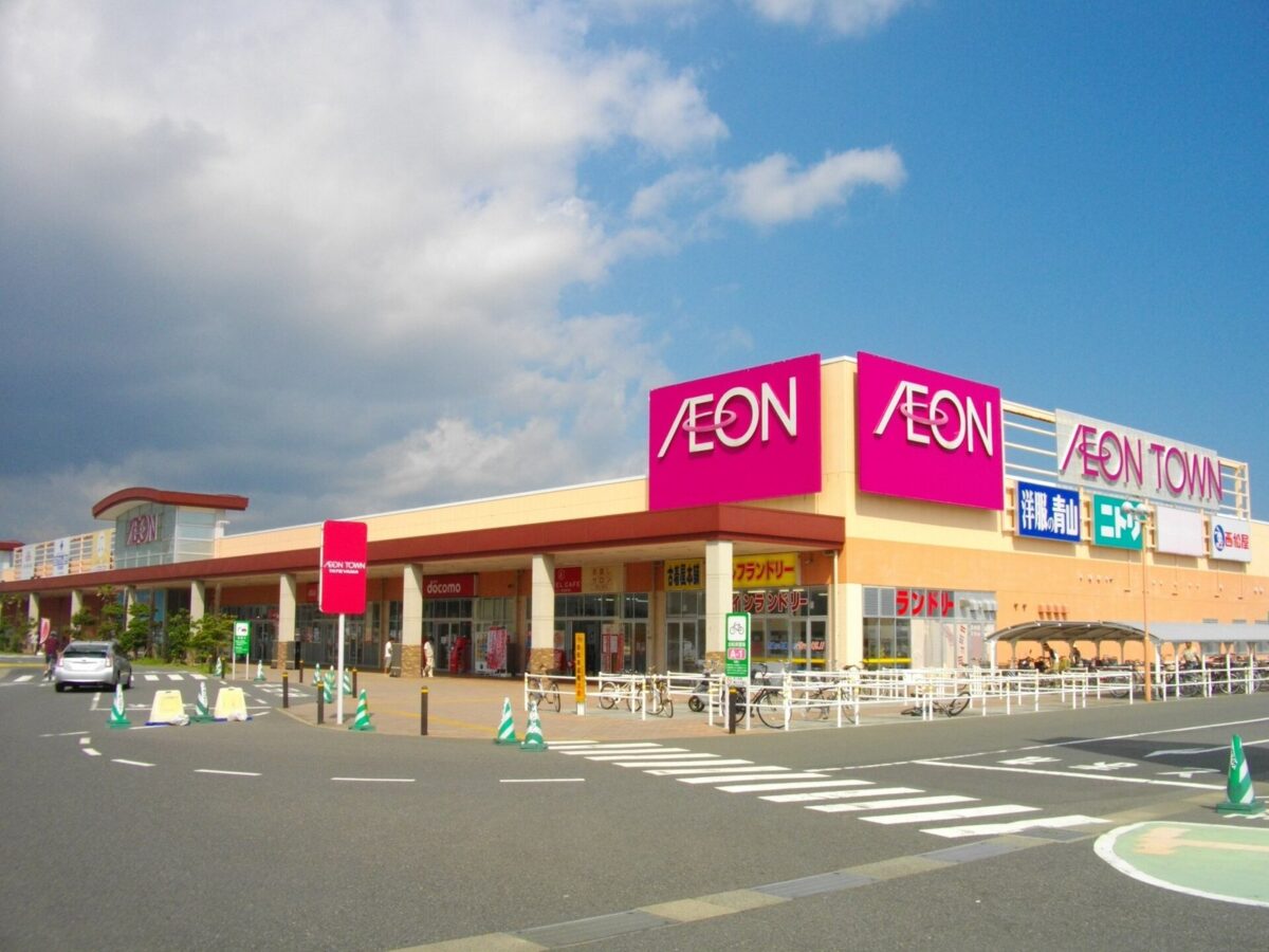 晴れた日のショッピングセンター。赤い「AEON」の看板の横に、緑の「ニトリ」の看板が並び、手前に広い道路と横断歩道がある
