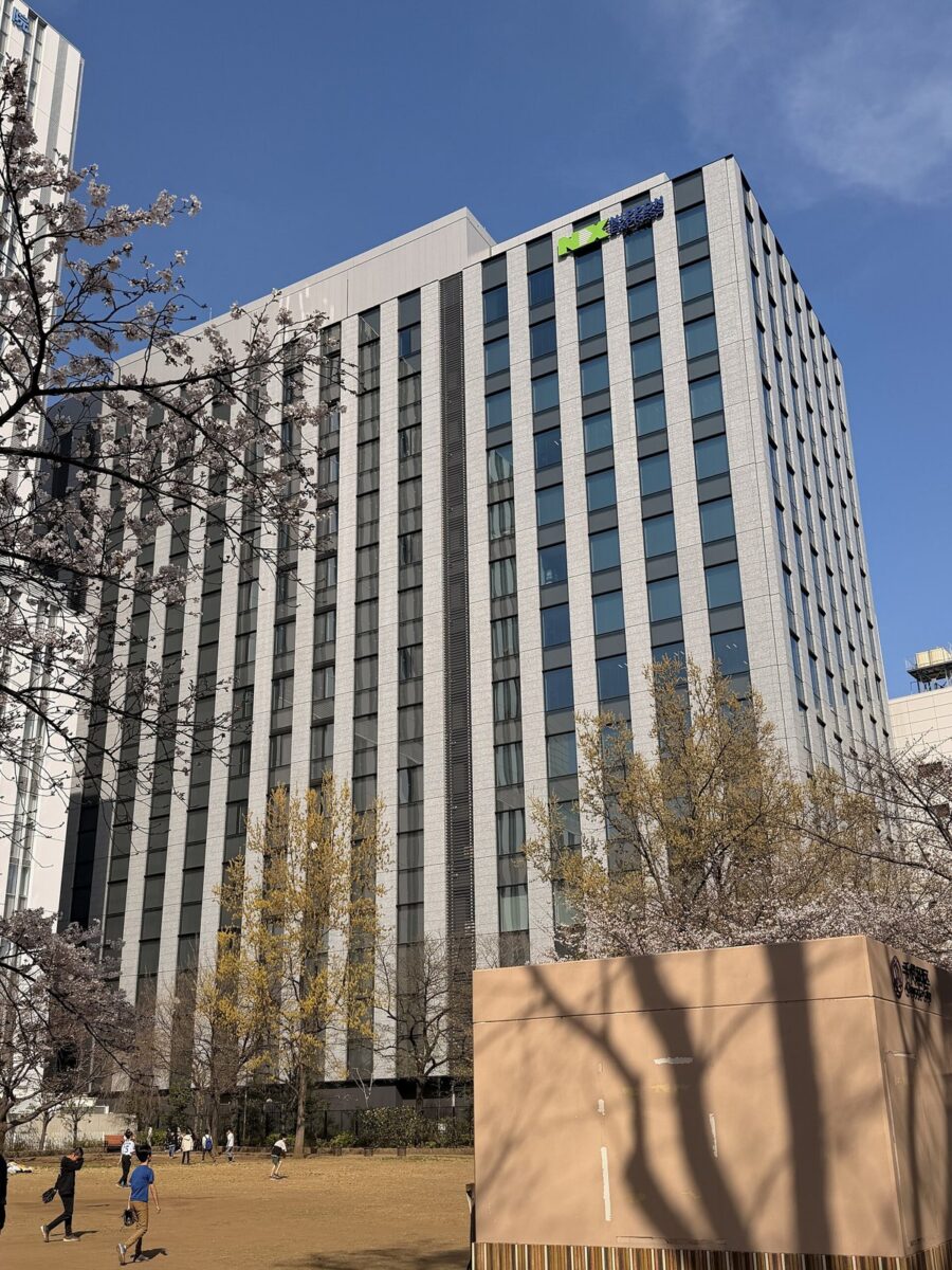 桜の咲く公園の向こうに建つ、灰色の縦じまの高いビル。屋上の近くに緑と青の「NX」のロゴ。NIPPON EXPRESSホールディングスと日本通運の本社