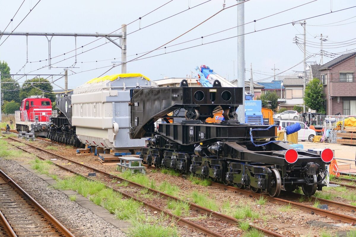 駅の線路に止まっている、たくさんの車輪がついた黒くて長い貨車。真ん中に灰色の大きな変圧器を挟み、後ろに赤い機関車がつながっている