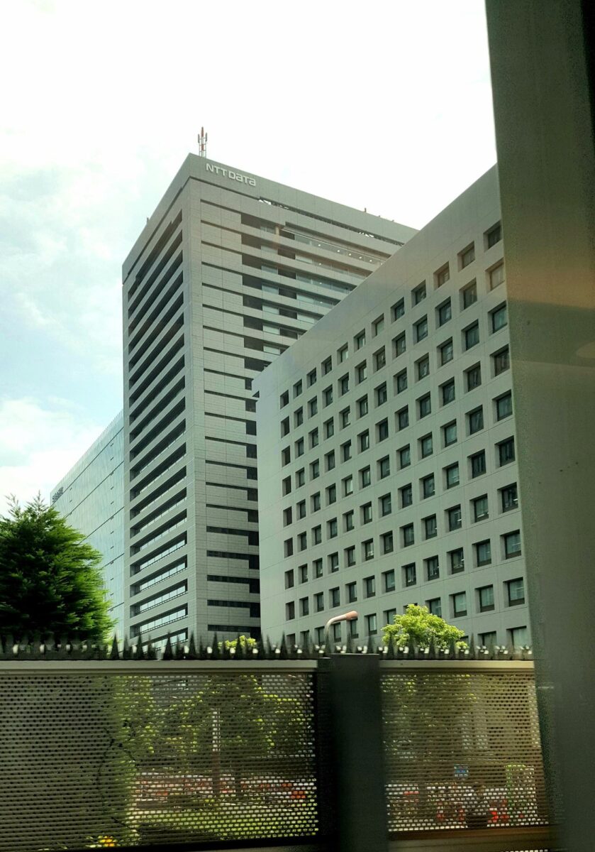 てっぺんに NTT DATA の文字がある灰色の高いビルと、窓が並ぶ低い別館。手前に木と柵がある