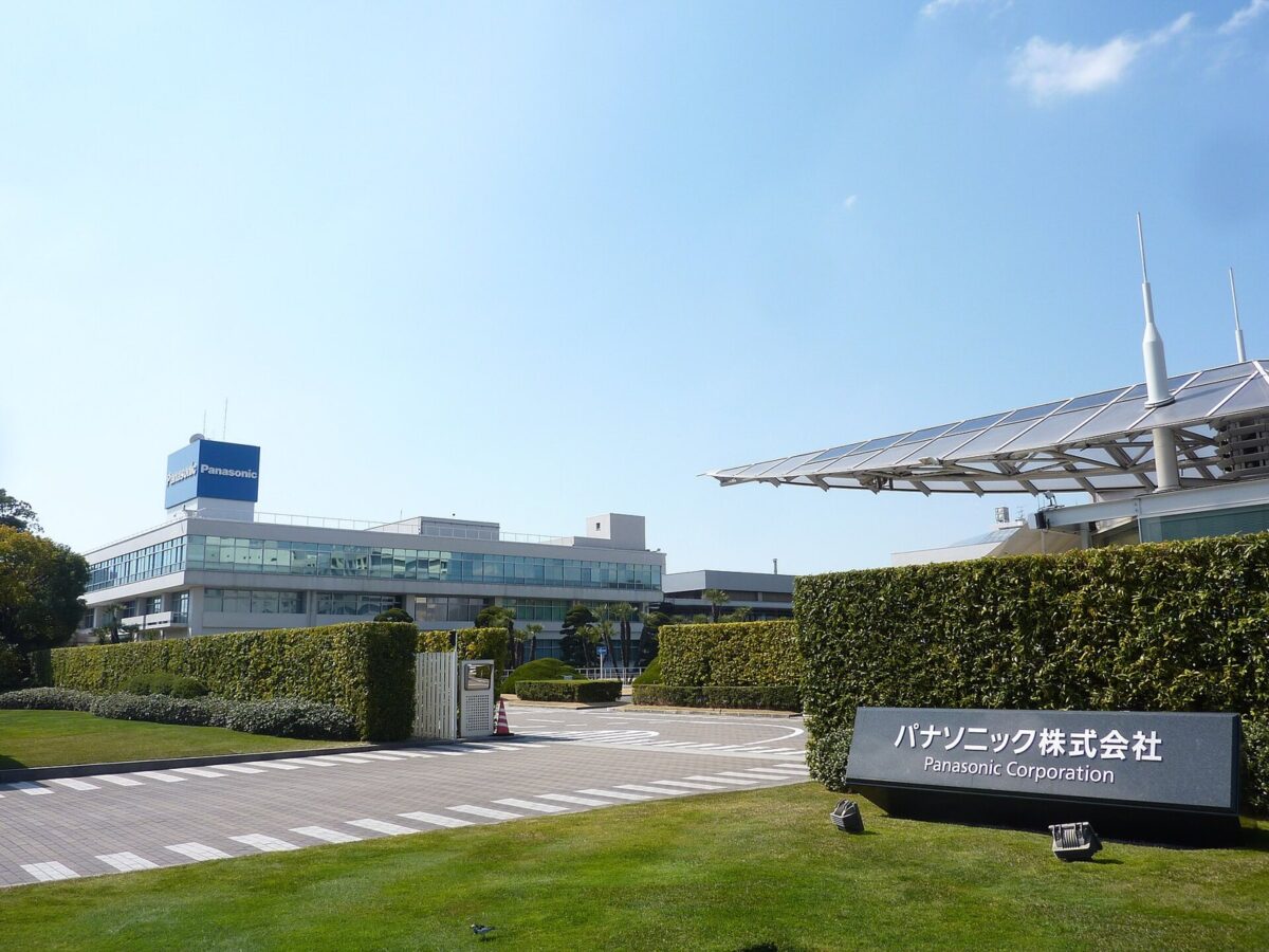 青空の下、芝生と生け垣の向こうに、青い看板のついた白い低い建物と、会社の名前の石の札が見える