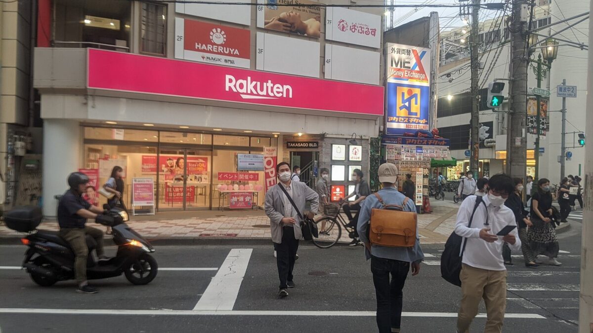 街の角にある、ピンクの「Rakuten」の看板のお店。お店の前の横断歩道を、人が渡っている
