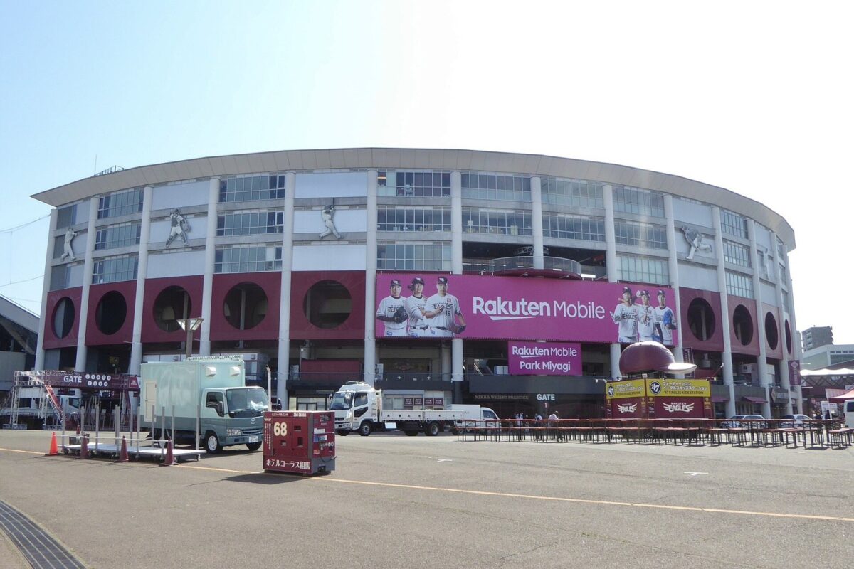 円い形の野球場の外壁に、赤い帯と「Rakuten Mobile」の文字と選手の大きな写真が並び、手前に広い広場がある