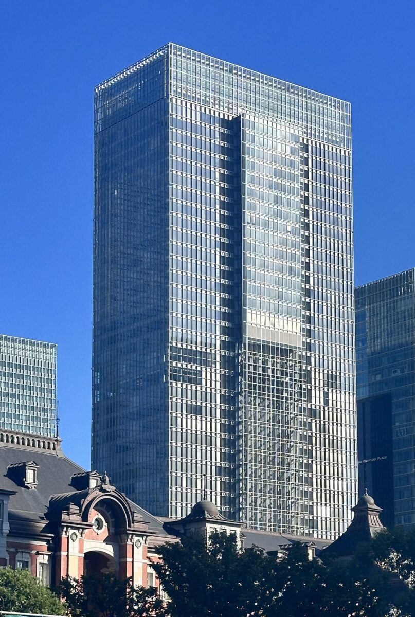 青空の下、赤レンガの東京駅の屋根の向こうに建つ、ガラス張りの高層ビルの写真