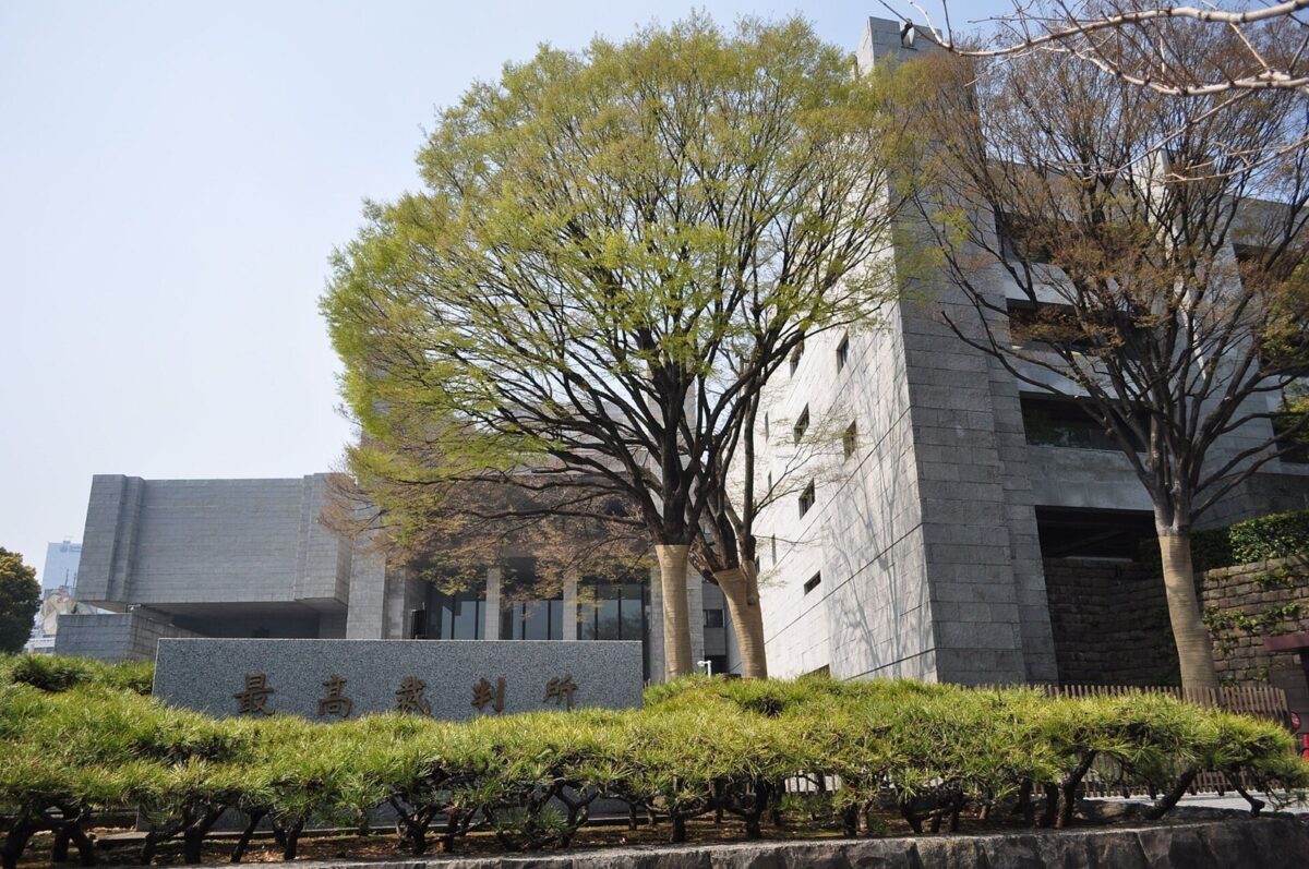 灰色の石の壁が角ばって重なる大きな建物。手前に大きな木と植え込み、「最高裁判所」と刻まれた石の門標がある