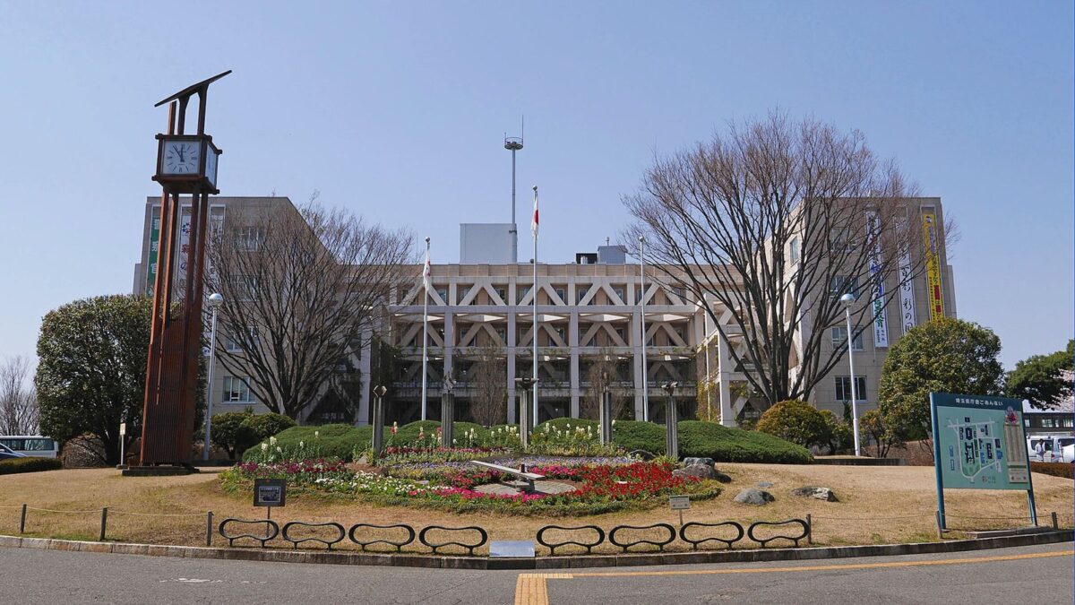 青空の下、中央に白い斜めの格子が並ぶ横長の庁舎。手前に丸い花壇と冬枯れの木々、左に時計塔