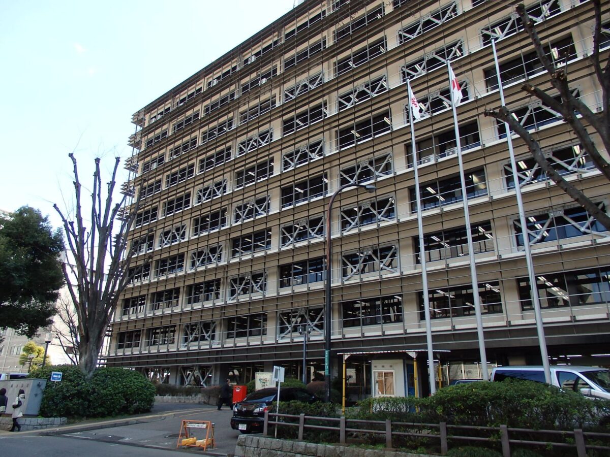 青空の下、横に長い灰色の高層の庁舎。壁一面に格子のような窓が並び、手前に木と植え込みと道路がある