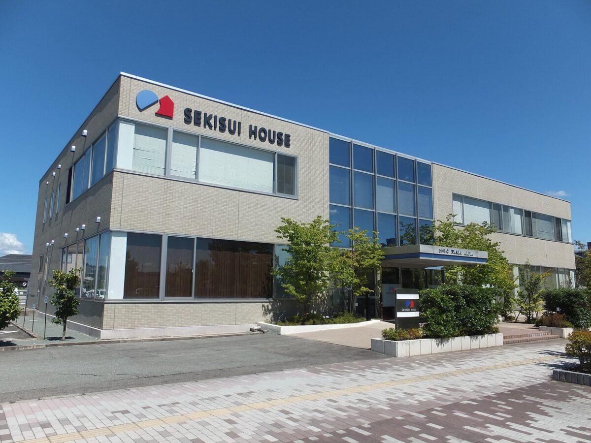 晴れた日の、灰色の外壁とガラス窓の2階建ての建物。入口の上に「SEKISUI HOUSE」の看板があり、手前に植え込みと石畳がある