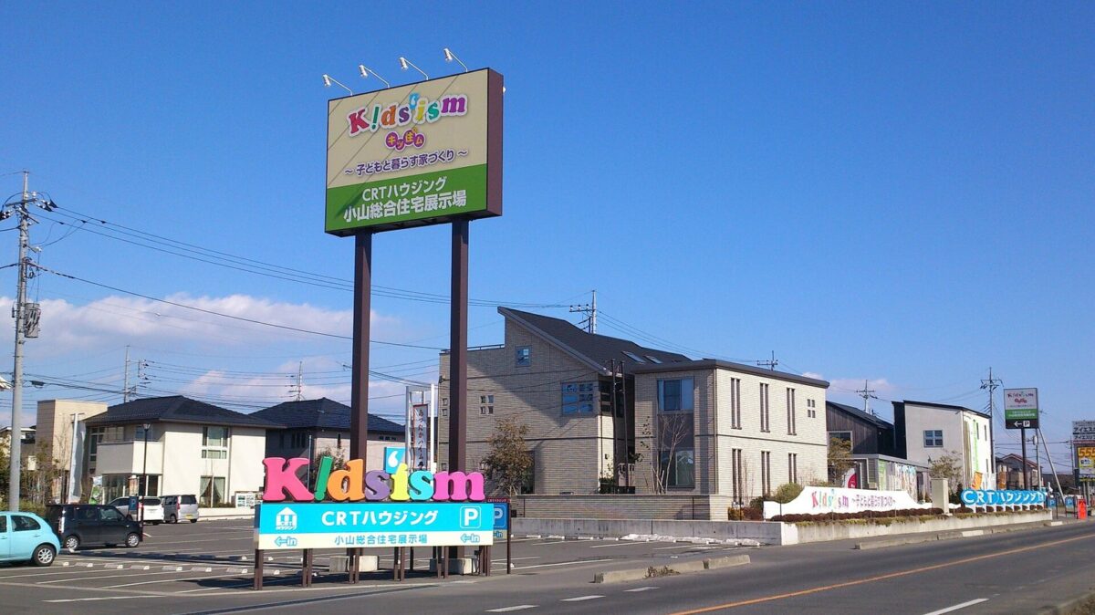 晴れた日の住宅展示場。高い看板の奥に、形や色の違う一戸建てのモデルハウスが並び、手前に道路と駐車場がある