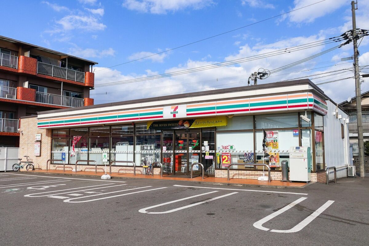青空の下、白い壁に緑・オレンジ・赤の帯が入ったセブン-イレブンの平屋の店があり、手前に白線を引いた広い駐車場が広がっている