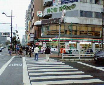 大きな交差点の角に建つビルの1階に、緑とオレンジの帯の看板のコンビニがあり、横断歩道を人が渡っている