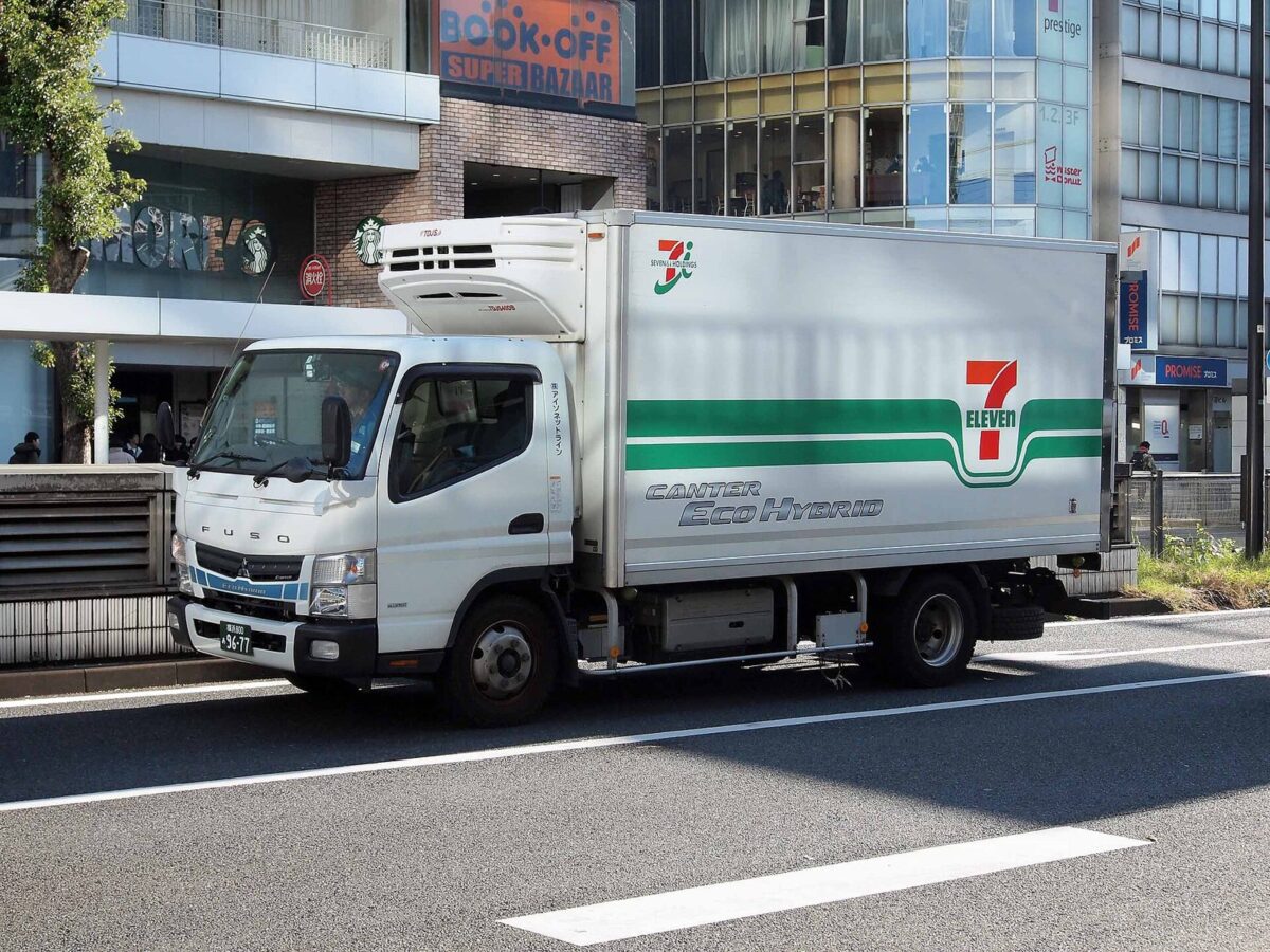 駅前の大通りに、荷台に緑の帯とセブン-イレブンのマークが入った白い冷蔵トラックが止まっている
