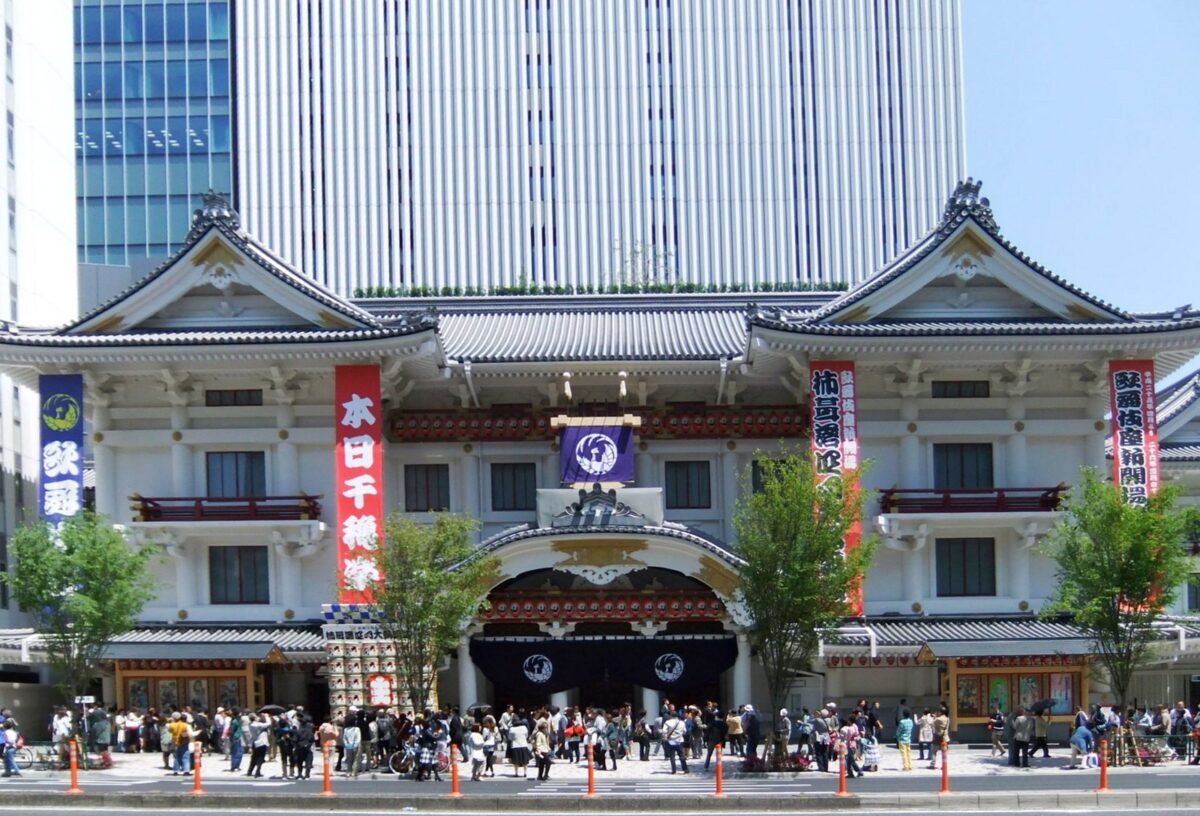 反り返った瓦屋根と白い壁の伝統的な劇場の正面に大勢の人が並び、その後ろに高層ビルがそびえる写真