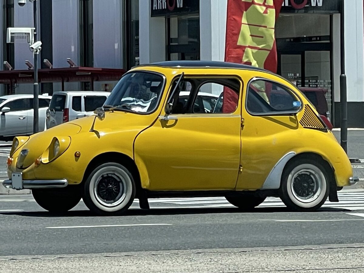 町の交差点を走る、黄色い丸い形の小さな古い車。屋根は黒く、白いふちのタイヤを付けている