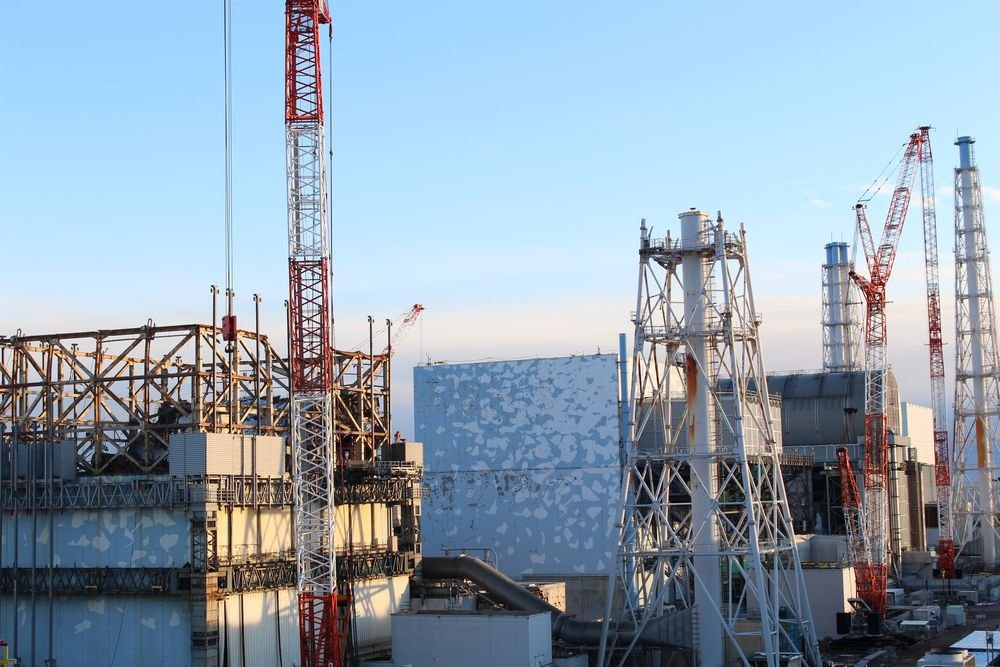 青空の下、鉄骨がむき出しの建物や、四角い覆いの建物が並び、その間に赤と白の大きなクレーンと鉄塔が何本も立っている