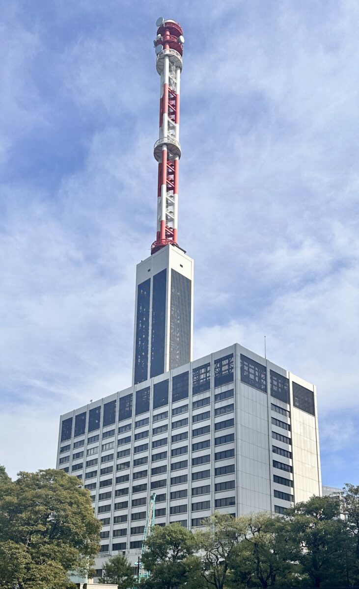 木々の向こうに立つ白い高層ビルと、その屋上から伸びる赤と白の高い鉄塔を見上げた写真