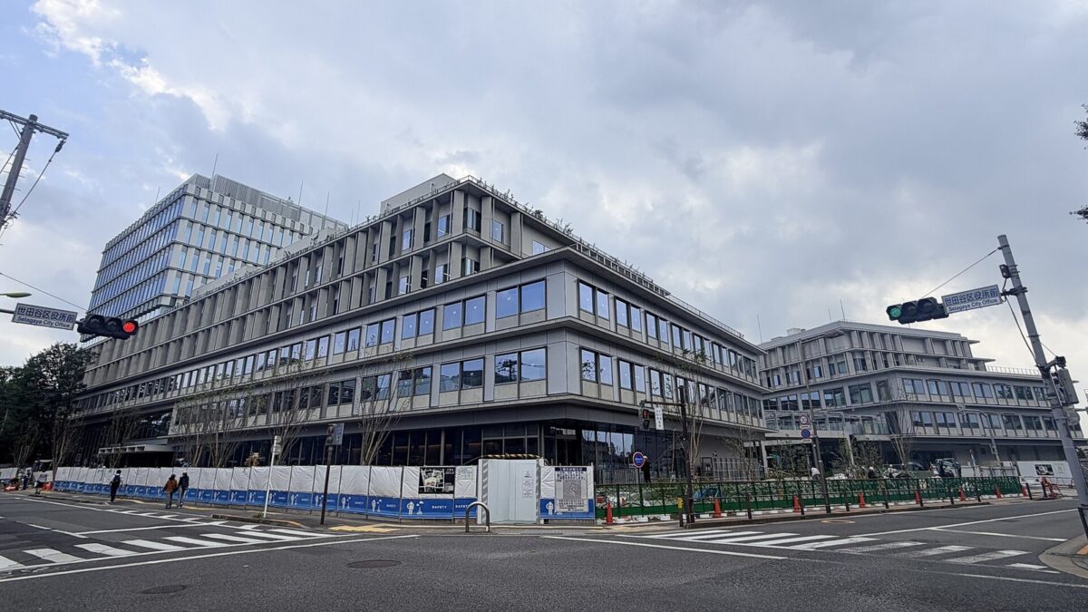 曇り空の下、交差点の角に建つ横に長い灰色の区役所の建物。手前に横断歩道と信号