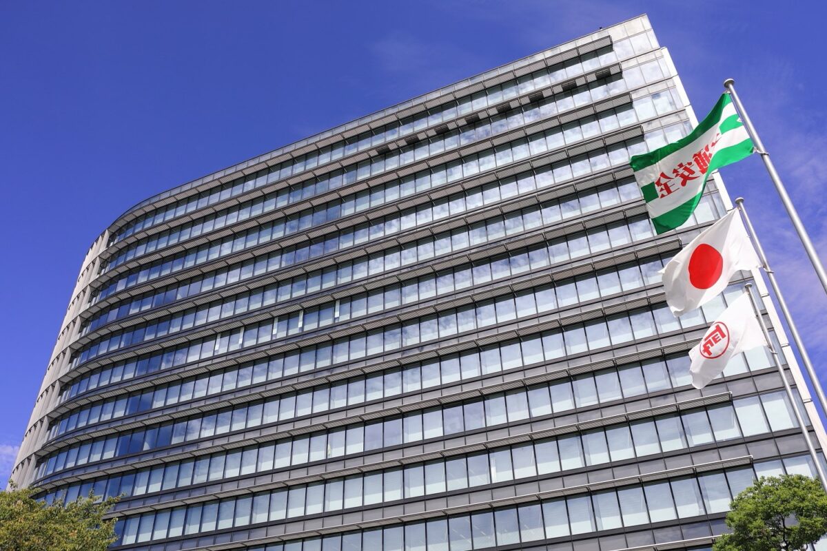 青空の下に建つ、ガラス窓が何段も並ぶ高いビル。右に日の丸などの旗が立っている
