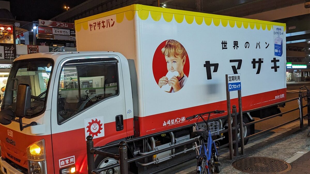 夜の町に止まった、白と赤のトラック。荷台に「世界のパン ヤマザキ」の文字と、パンを食べる女の子の絵が描かれている