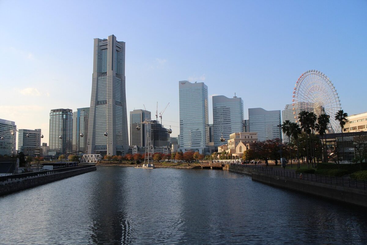 青空の下、運河の向こうに先のとがった高い超高層ビルとガラスのオフィスビルが並び、右に大きな観覧車とヤシの木が見える